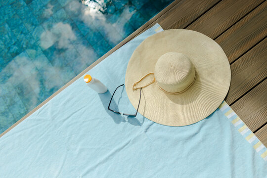 Woven straw sun hat, black sunglasses and sunscreen are resting on light blue towel by pool