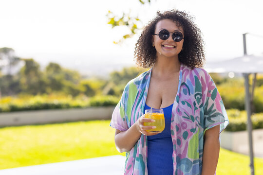 Woman standing on sunlit patio wearing blue dress coverup holding glass orange-juice-straw