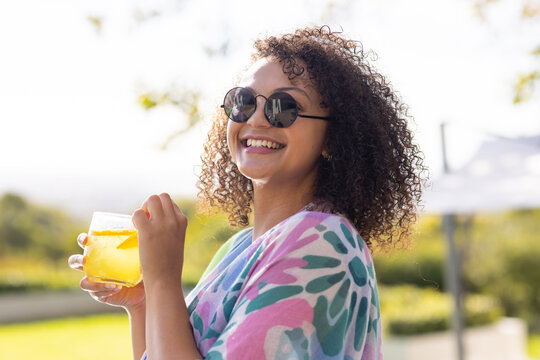 Adult African American woman standing and smiling on sunny terrace, holding glass with lemon wedge