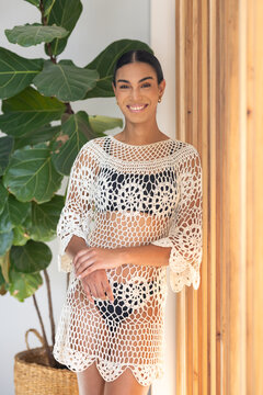 Woman standing smiling, wearing white crochet dress over black bikini and gold hoops in studio