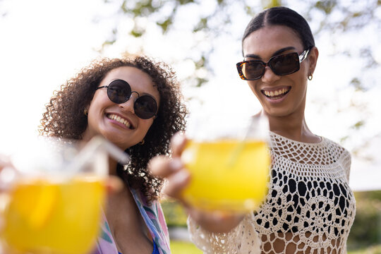 African American female friends toasting with glasses and clear straws at park in swimwear, shades