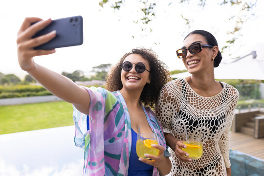 African American women friends posing with phone and drinks in swimwear on pool deck