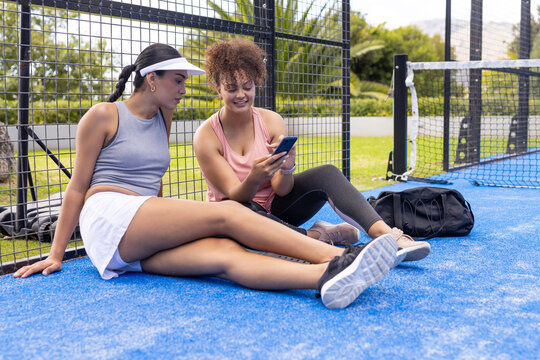 Diverse female friends sitting on blue court in sportswear, looking at smartphone near duffel