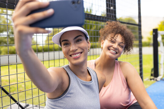 African American friends sitting at outdoor court taking smartphone selfie, wearing visor and tanks