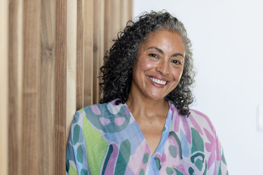 Senior woman leaning on wooden slat wall in studio smiling wearing colorful top and stud earrings