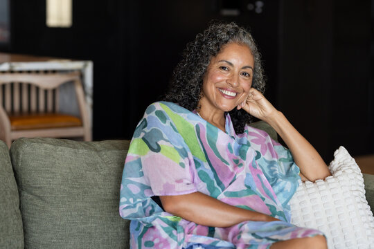 Senior African American woman sitting on green sofa at home wearing colorful kaftan and smiling