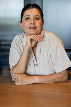 Female sitting at wooden table in office, resting chin on right hand, wearing beige shirt, ring