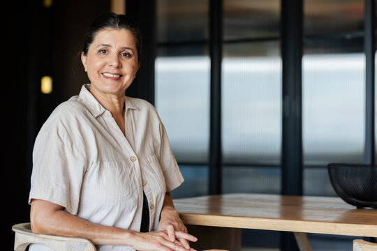 Senior woman sitting at wooden table in modern office lounge wearing beige shirt, black basket near