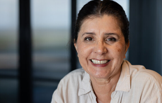 Mature woman sitting and smiling, facing camera by glass panels wearing light collared shirt