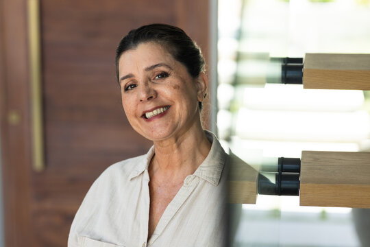 Mature adult female leaning on glass partition, smiling in light beige shirt near floating stairs