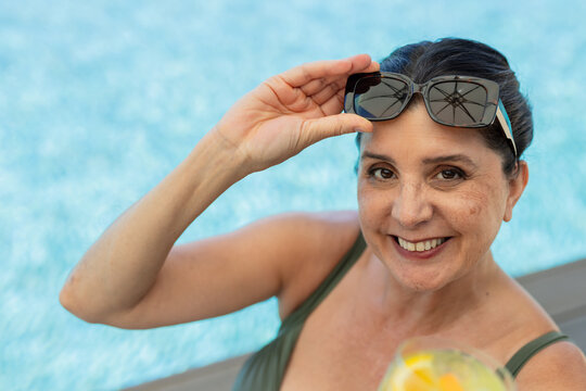 Mature woman lifting sunglasses, holding citrus drink at pool edge in olive-green swimsuit