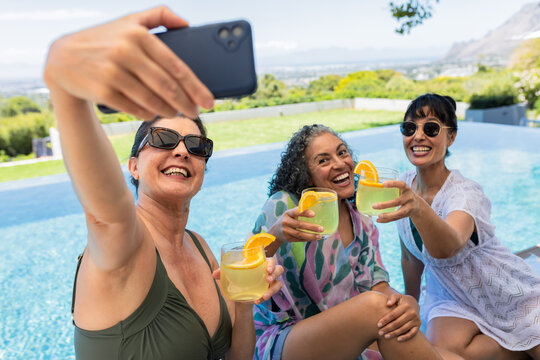 Diverse female friends sitting at pool taking selfie with phone holding cocktails with sunglasses