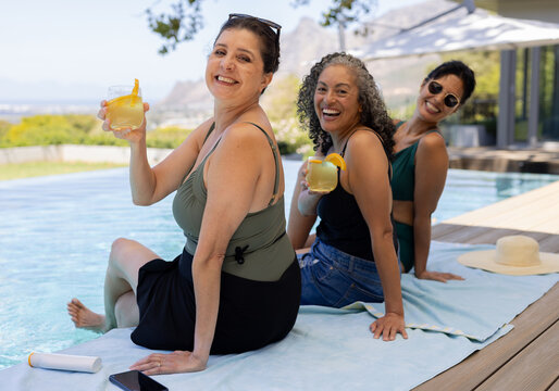 Diverse female friends sitting at infinity pool deck holding citrus drinks phone and SPF beside hat