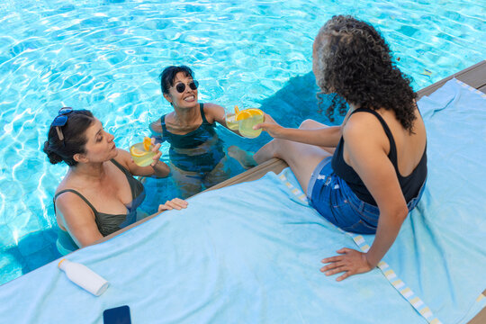 Female friends in pool wearing swimsuits, holding yellow cups, one sitting reaching from blue towel