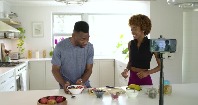 Diverse co-hosts responding to smartphone tripod preparing and presenting fruit bowl in kitchen