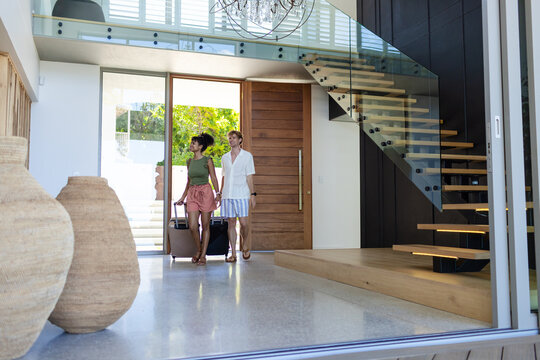 Couple holding hands walking into home entry, pulling wheeled suitcases past woven vases