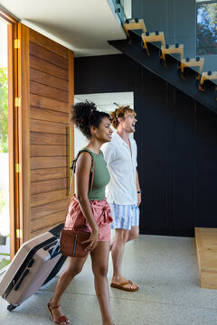 Diverse couple entering modern home, pulling pale rolling suitcase under wooden pivot door