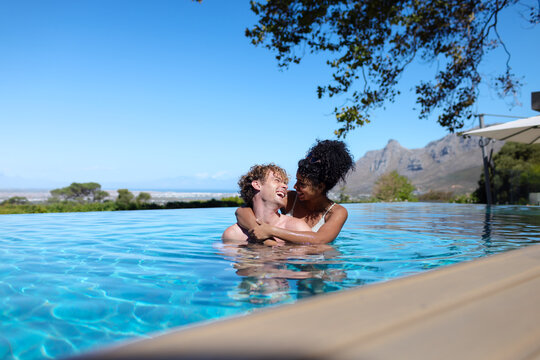 Couple in mid twenties wearing swimsuits embracing and laughing in infinity pool near wooden deck