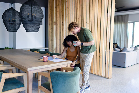 Couple leaning over paperwork at open-plan dining table with mugs and green chairs, copy space