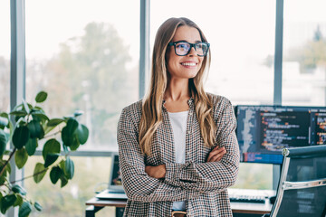 Naklejka premium Confident young female software developer smiling with arms crossed in a bright modern office in front of monitors displaying code