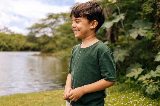 Brazilian boy smiling near a calm lake surrounded by greenery, portraying joyful childhood, outdoor freedom and connection with nature in a peaceful environment.
