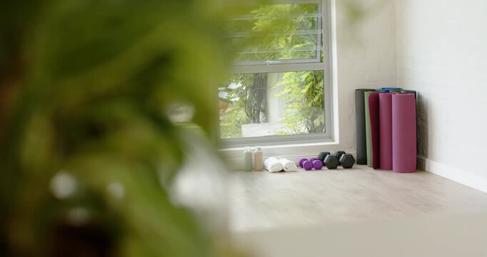Camera is shifting focus, revealing stacked yoga mats, dumbbells and water bottle for home workout