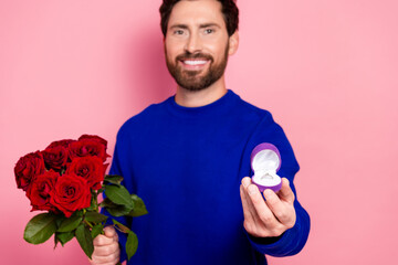 Young man in blue sweater offers red roses and engagement ring in pink studio setting