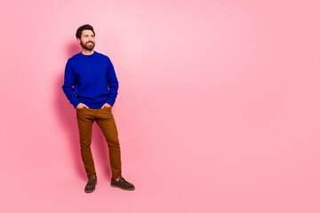 Man in blue sweater and brown pants stands smiling against pink background