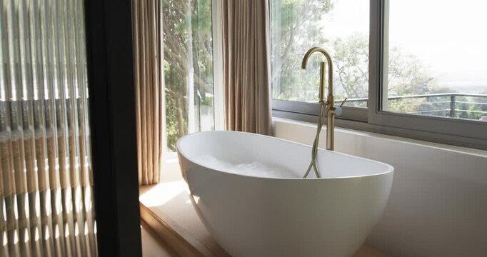 Camera is panning right, revealing bubbly standalone white oval tub and brass faucet to show window