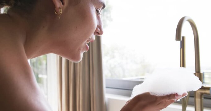 Woman holding bath foam at bathtub then blowing and watching foam drifting toward brass faucet