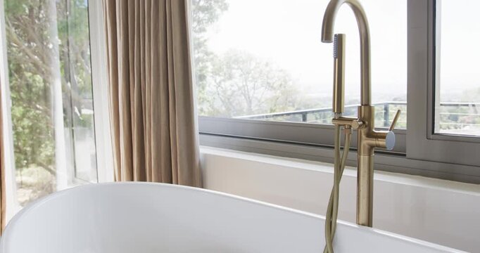 Off-screen faucet is turning on, pouring water into tub and forming foam, preparing bath by window