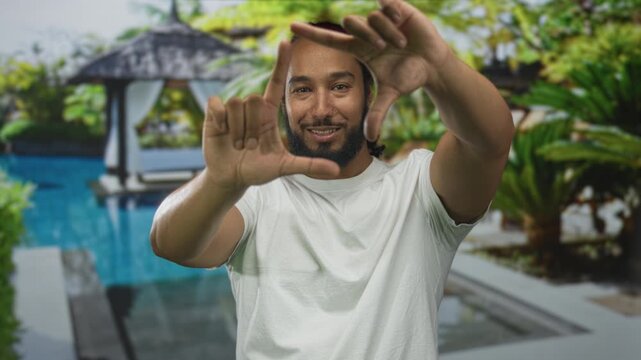 Man framing with hands at a resort pool outdoors, smiling and making a rectangular frame gesture with fingers while wearing a white t shirt; calm vacation moment.