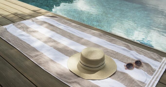 Beige towel with straw hat and sunglasses is resting on deck while breeze is rippling pool