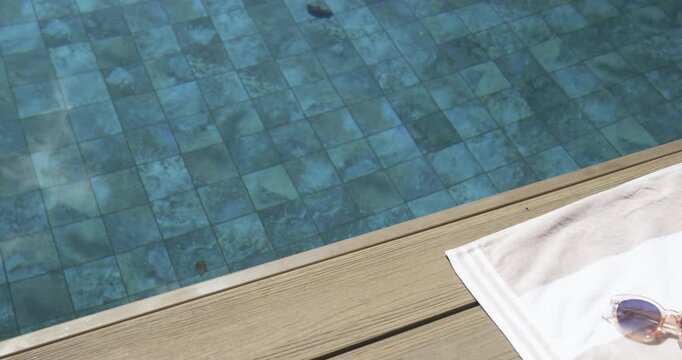 Camera is shifting right to reveal white towel with round sunglasses while pool water is rippling
