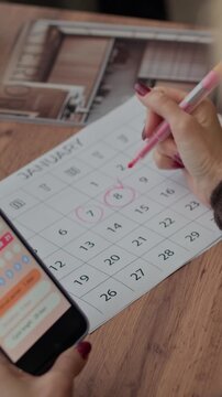 Vertical shot of unrecognizable woman checking menstrual period on smartphone and highlighting dates in calendar while sitting at home table