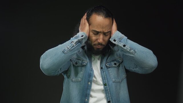 Man wearing denim jacket with hands covering ears and pained expression on black studio backdrop; stress headache recovery.