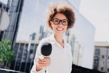 Confident young female reporter smiling with microphone in busy city street outside modern glass...