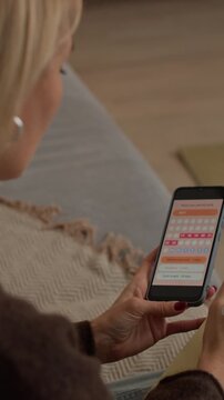 Vertical shot of middle-aged Caucasian woman checking menstrual cycle on smartphone and taking notes while sitting on sofa in living room