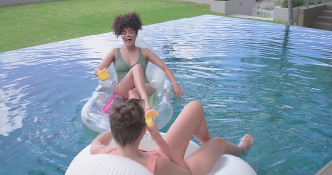 Warm sun urging diverse female friends on floats in infinity pool, holding yellow cups and laughing