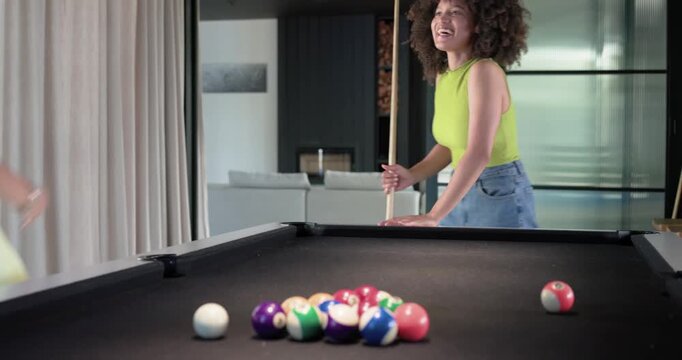 Diverse female pals curly friend breaking leading romper friend shooting at home pool table for fun