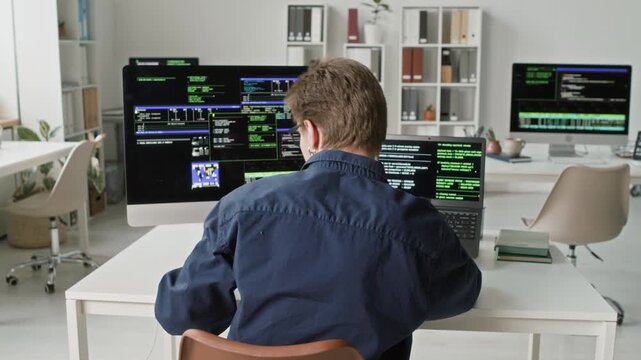 Rear view of young adult software male developer typing on computer and laptop using programming language while fixing mistakes in code at workstation in modern IT company office