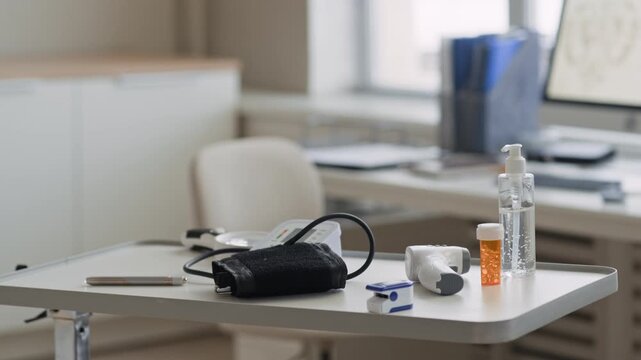 Indoor eye level close up broll shot of medical examination table with blood pressure monitor, sanitizer bottle, pill container, thermometer, and diagnostic tools in clean and sterile clinic office