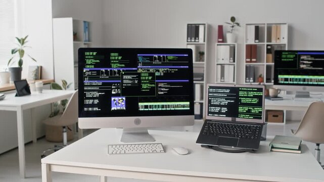 No people shot of coding setup with desktop computer and laptop displaying lines of code placed on programmers desk at modern open plan office