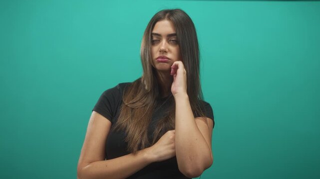 Woman touching chin with hand and arm folded holding elbow in studio wearing black shirt against teal backdrop; pensive.
