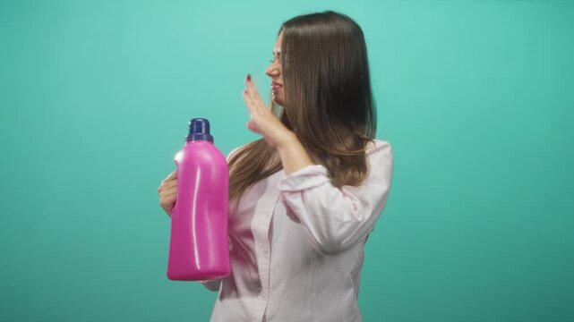 Young hispanic woman holds bright pink detergent bottle, pinches nose and turns away in studio; disgust caution.