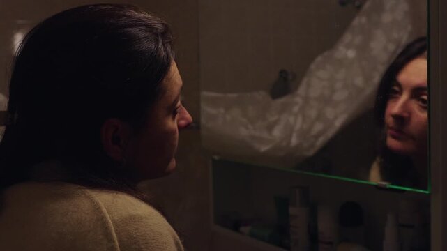 Middle aged woman in 40s looking into mirror with resigned expression, intimate moment of self reflection about aging and life transition