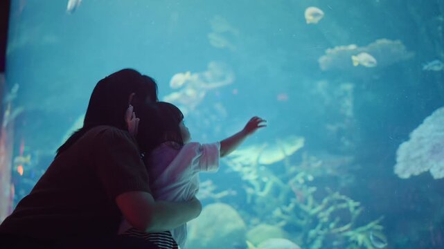 Back view Asian mother and her toddler daughter standing in front of a large aquarium tank, where the curious child is reaching out her hand toward a swimming fish.