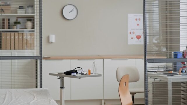 Indoor wide eye level establishing shot of empty medical examination room with exam bed, adjustable table holding medical devices, chair, wall clock and anatomy posters in clinical healthcare setting
