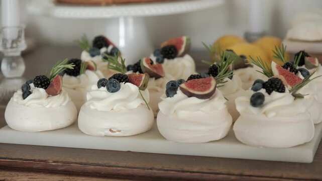 A row of mini pavlova cakes topped with whipped cream, figs, blackberries, blueberries, and rosemary on a marble platter