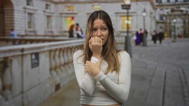 Woman bites nails with hand to chest on crowded city street beside stone building and lamppost; anxiety vulnerability.
